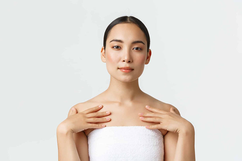 Head and shoulders of an Asian woman wearing a towel around her and her hands touching her collar bone. She has dark hair tied back and is looking directly at the camera.