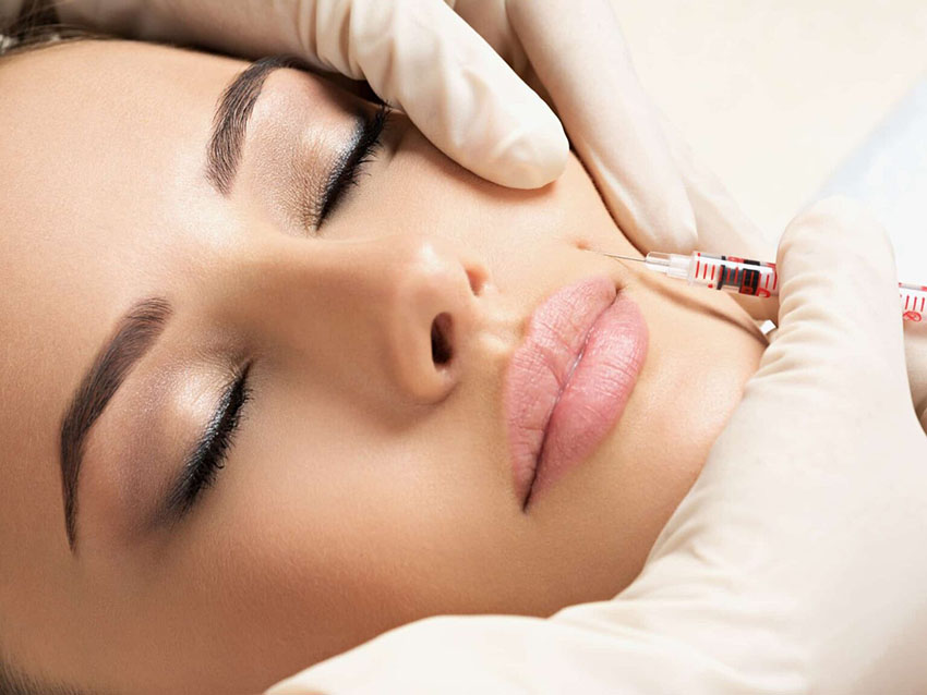Close up of a woman's face being held by a paid of white-gloved hands with an ultra-fine needle being held to the right and slightly above of the lips, pressing lightly on the skin.