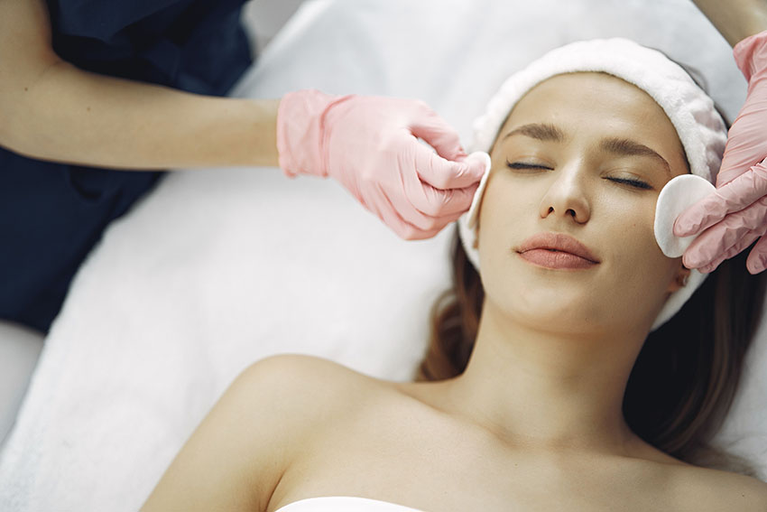 A close up of a woman lying on a treatment bed with her eyes closed, wearing a white headband and another person's hand in pink latex gloves stroking her cheek with a cotton wool pad.