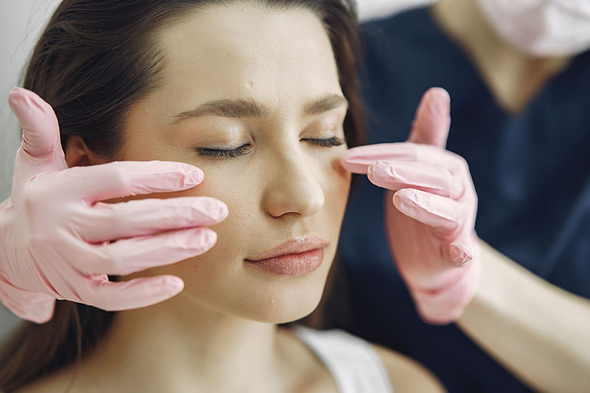 Close up of a woman's face with here eyes closed and a pair of hands wearing pink latex gloves touching her cheek bones.