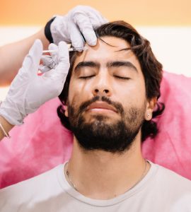 Head and shoulders of a man wearing a white t-shirt lying down with his eyes closed, being injected with anti-wrinkle treatment in his forehead with a fine needle by a someone wearing white latex gloves.
