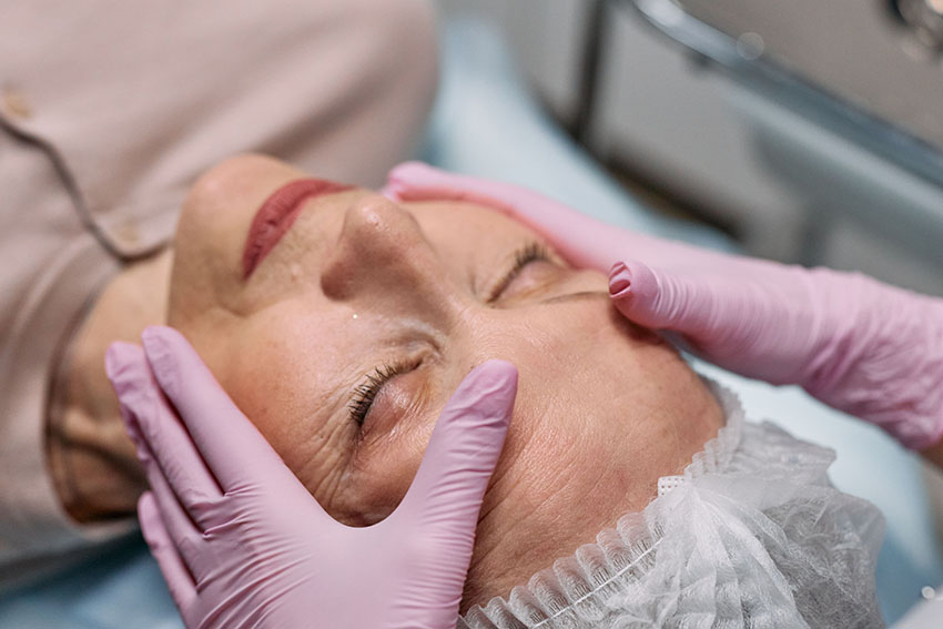 A close up of a mature woman's face with her eyes closed and someone else's hands wearing pink latex gloves touching her face in preparation for Morpheus8 Skin Tightening treatment.
