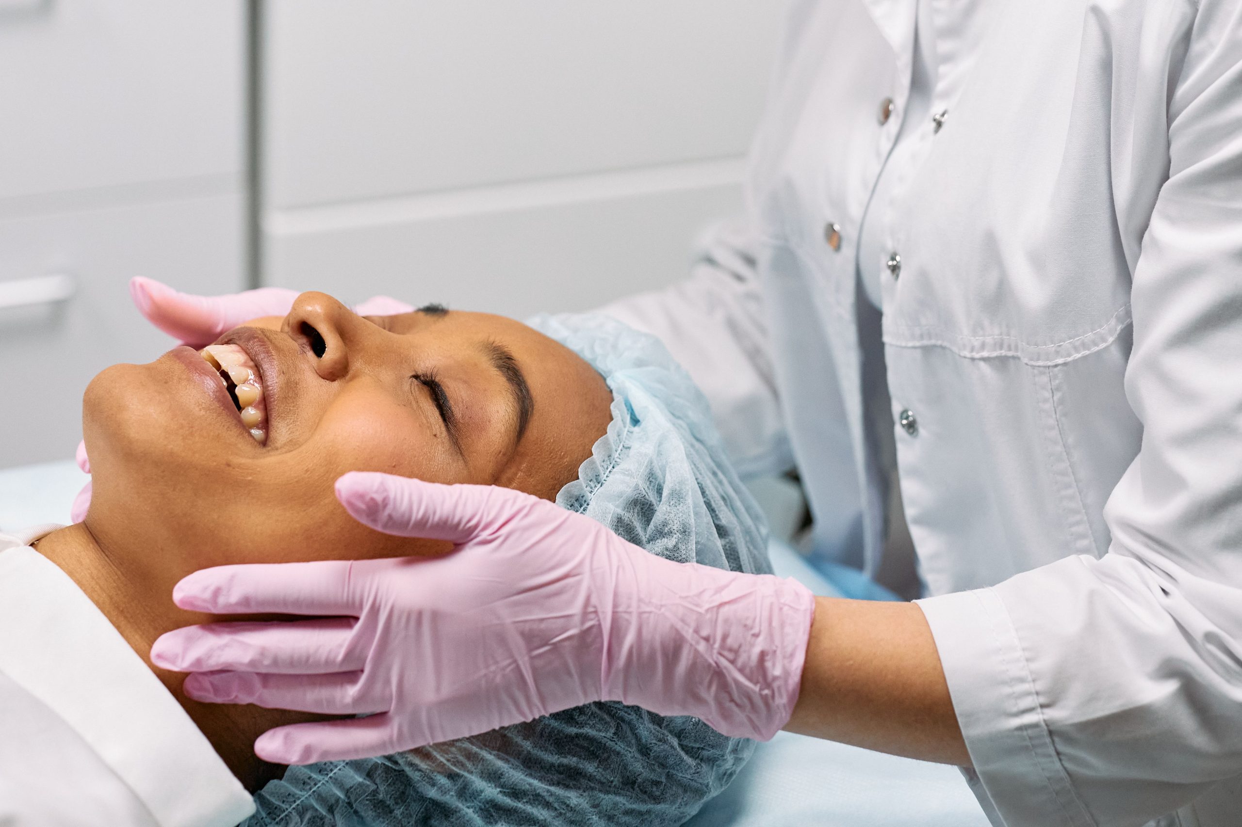 Head and shoulders of a smiling woman with her eyes closed lying on a treatment table with another person standing behind wearing a white doctors coat and pink latex gloves holding her face.