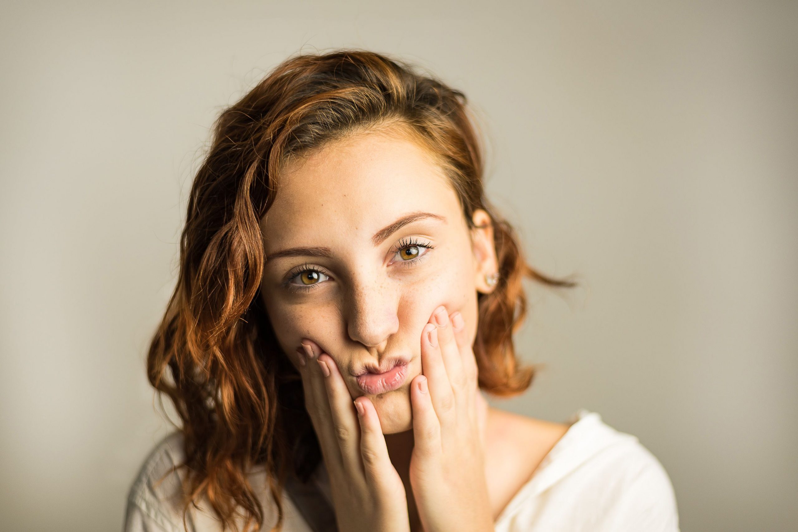 Head and shoulders of a woman with brown shoulder-length hair squishing her cheeks up with both hands on her face.