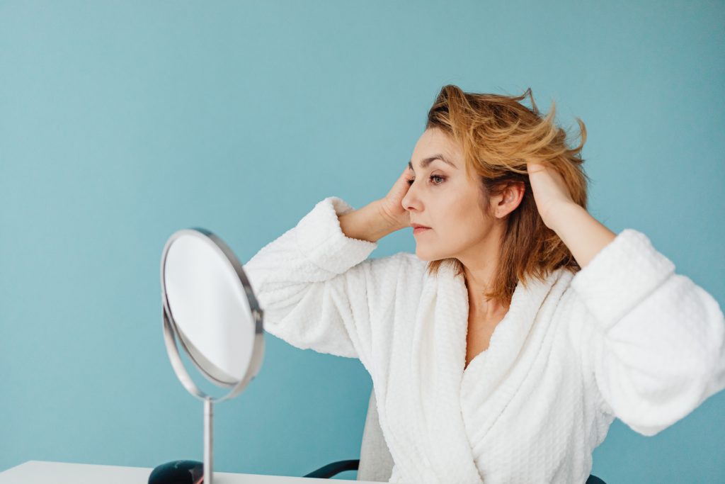 Woman wearing a white towelling robe with her hands touching her temples, looking in a small round mirror.