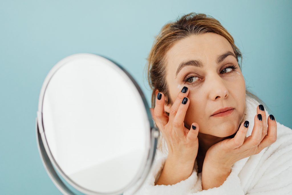 Close up of a woman looking in a round make-up mirror, with both hands lifted up to her face, one touching the skin just beneath her eye.
