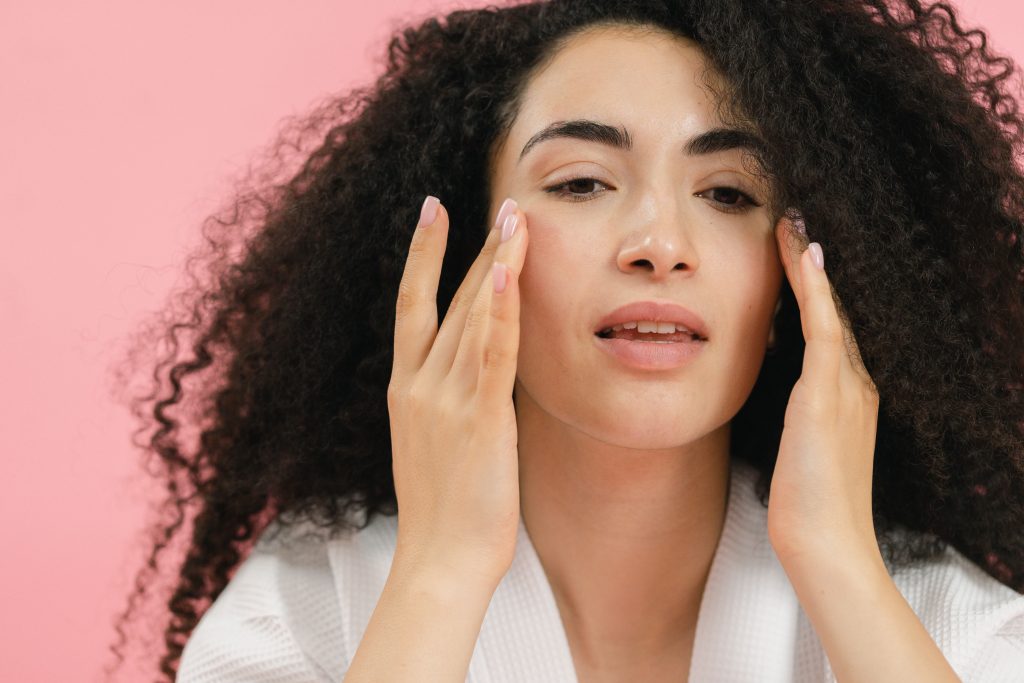 Close up of a woman's head and shoulders against a pink background. The woman has long dark curly hair and is touching her face with both hands on her cheeks.