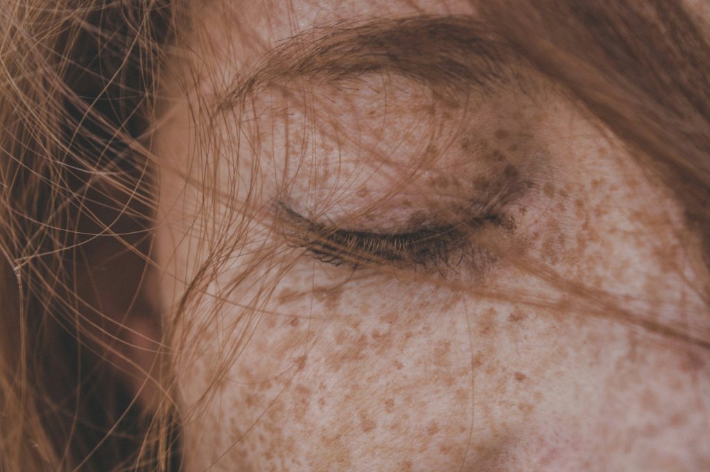 Close up of a woman's face showing one closed eye and eyebrow with strands of hair across the face and lots of freckles.